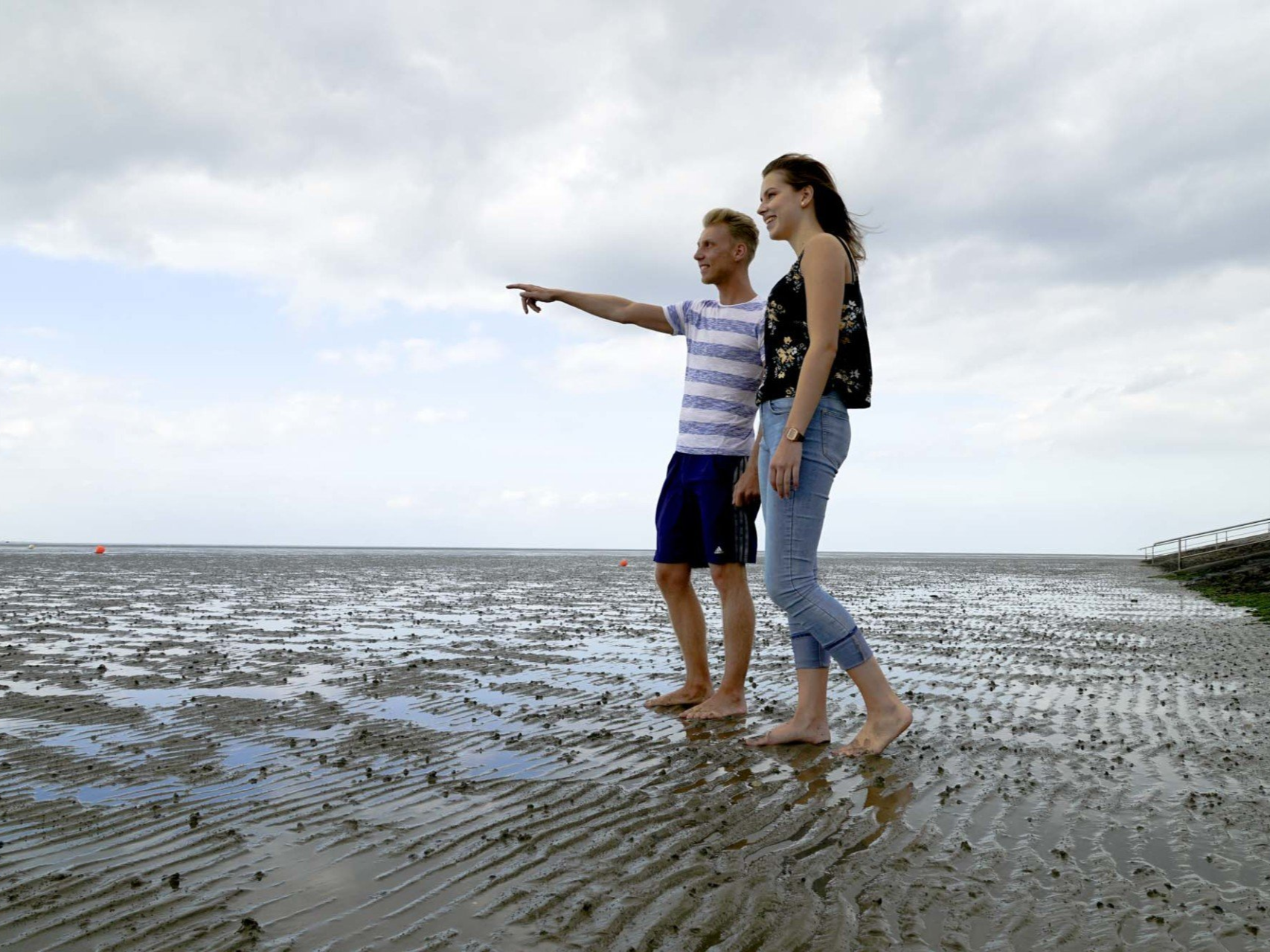 Zwei Personen stehen im nassen Sand, eine zeigt mit dem Finger. Bewölkter Himmel, Küstenlandschaft.
