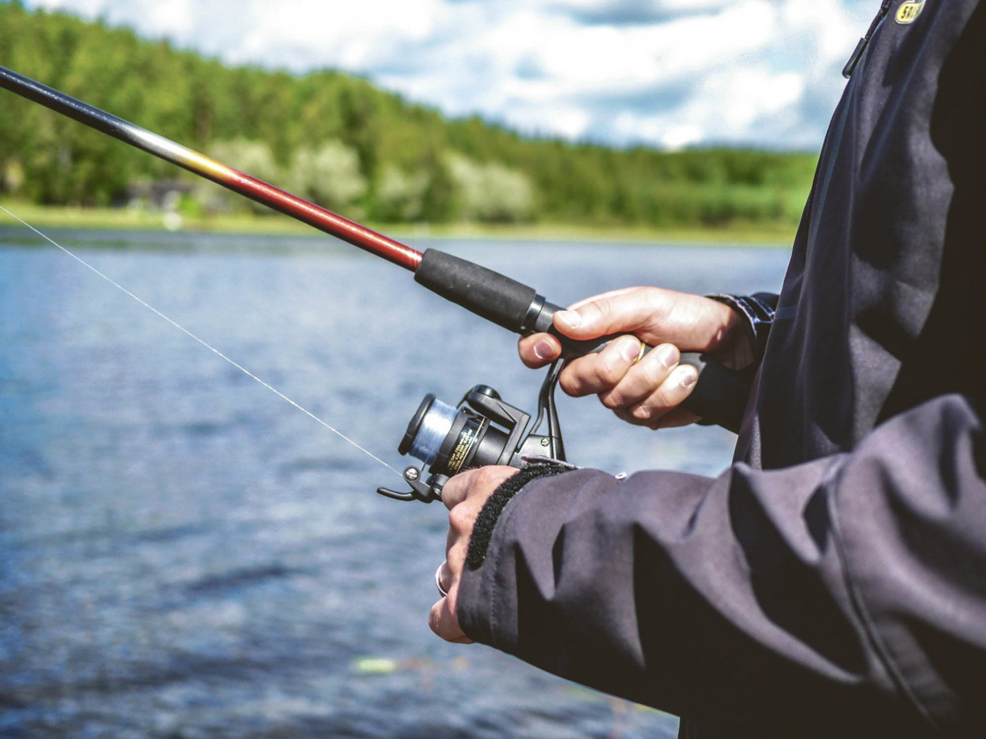Eine Person angelt und hält eine Angelrute über einen See. Im Hintergrund ist ein Wald zu sehen.