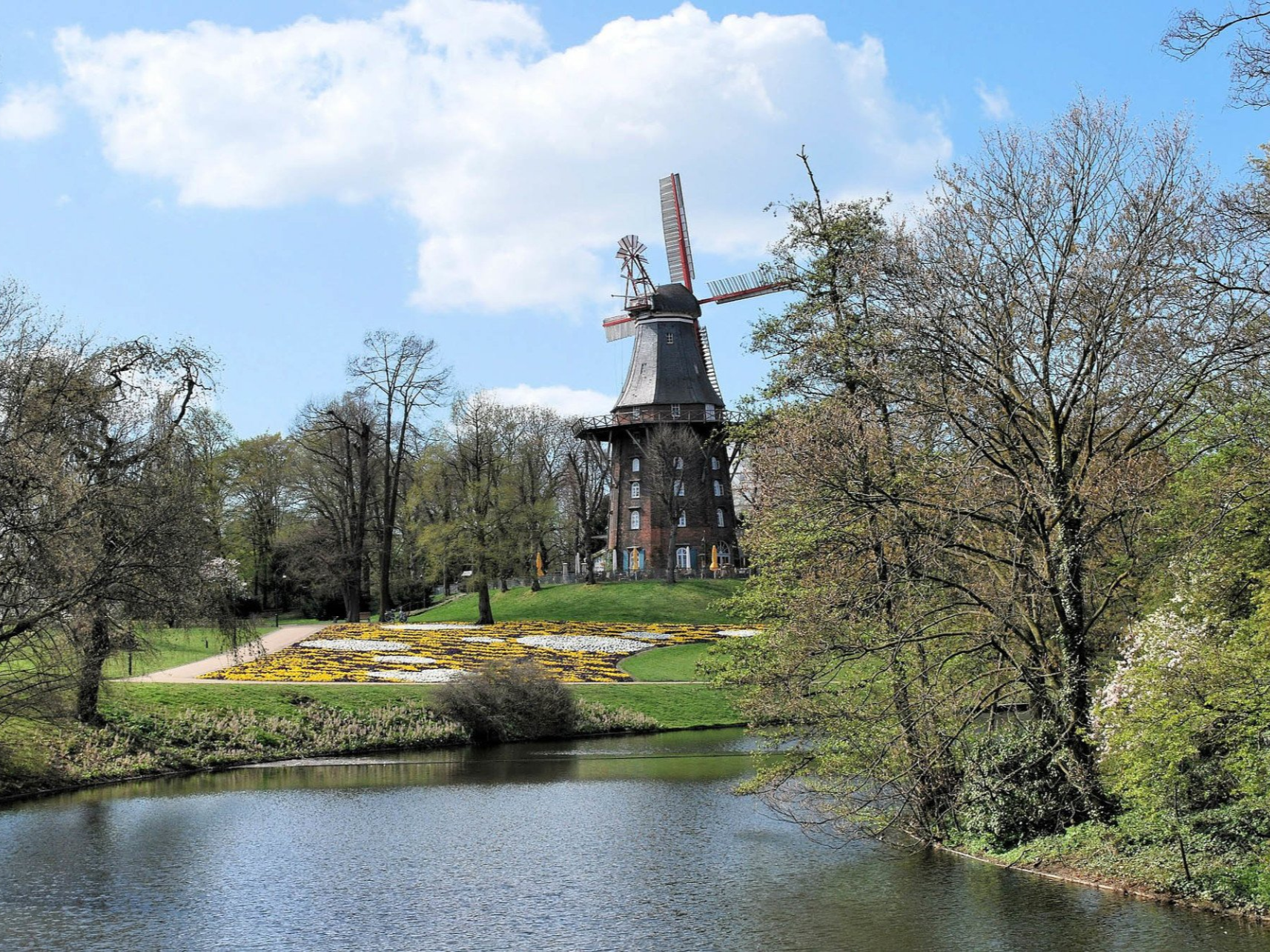 Eine Windmühle an einem Teich, umgeben von Bäumen und einer grünen Rasenfläche unter blauem Himmel.