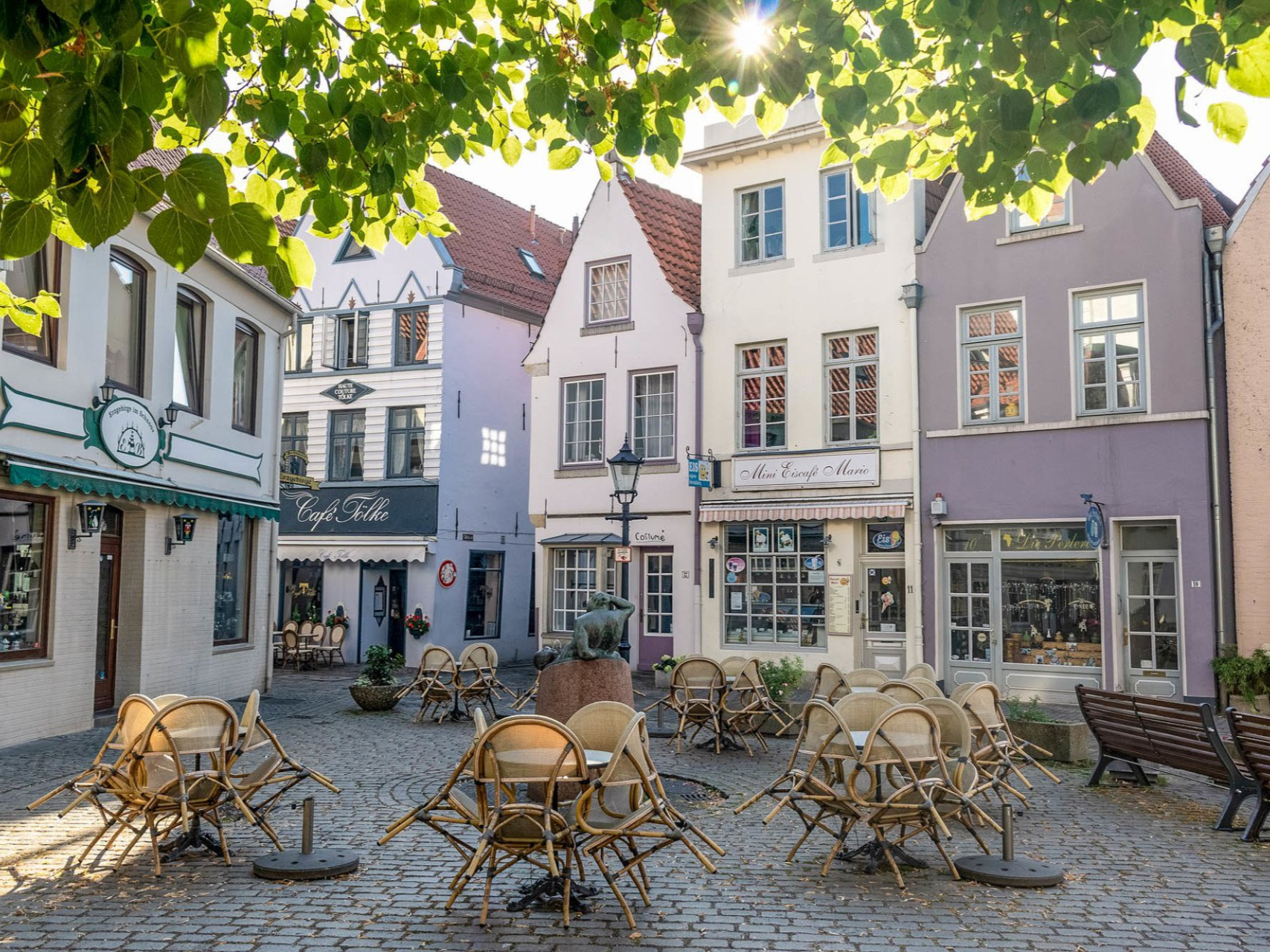 Kopfsteinpflasterplatz mit überdachten Außensitzplätzen eines Cafés, umgeben von farbenfrohen Gebäuden, sonniger Tag.