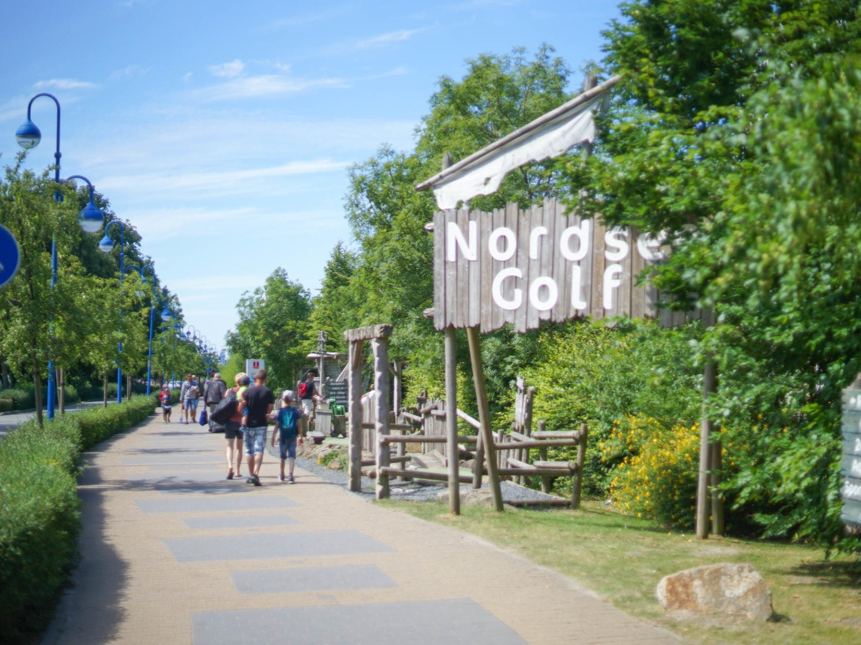 Gehweg mit Passanten, rechts ein Schild für Nordsee Golf, Bäume, blauer Himmel.
