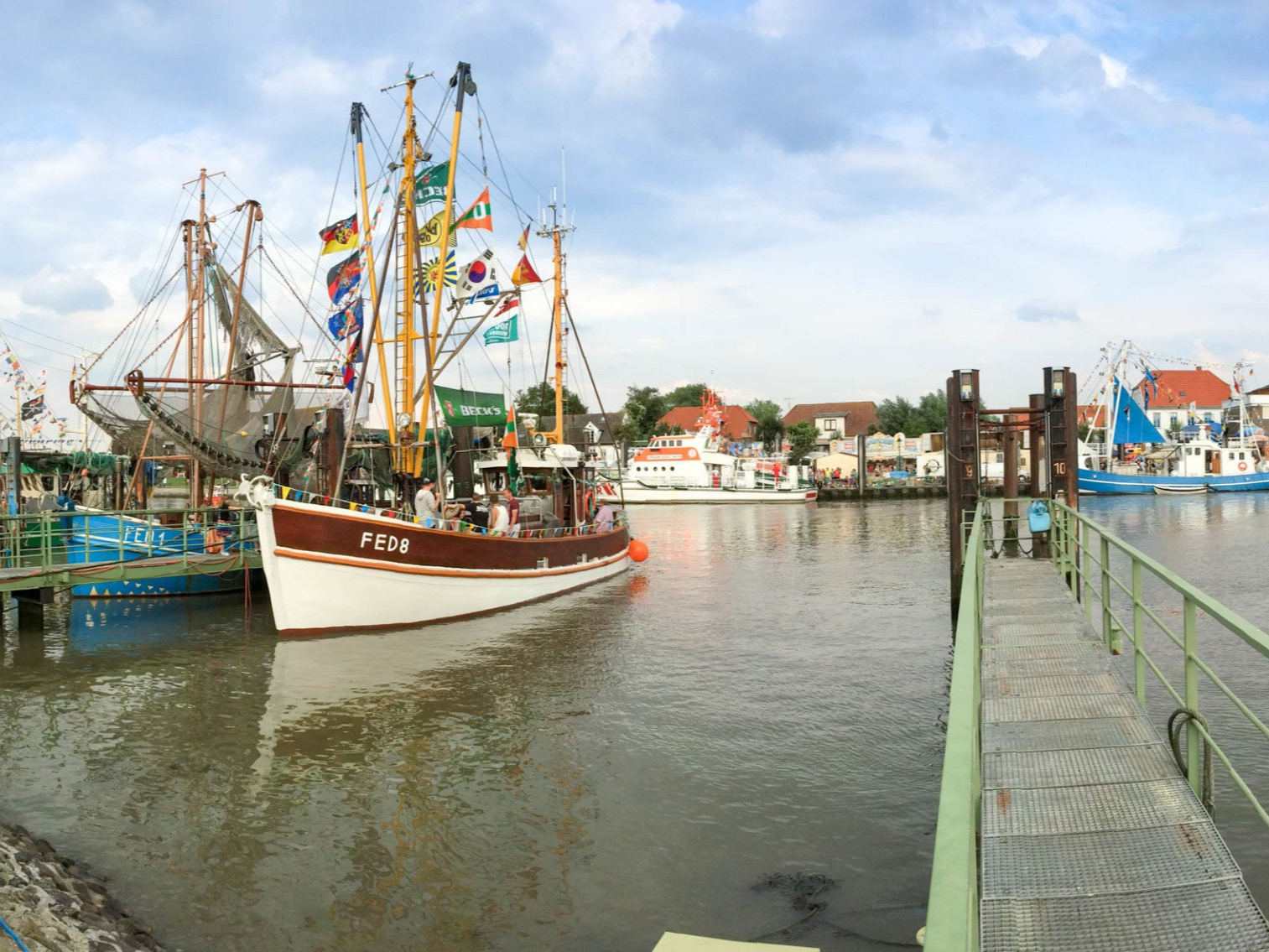 Fischerboote liegen im Hafen vor Anker, ein braun-weißes Boot im Fokus, Flaggen an den Masten, bewölkter Himmel.