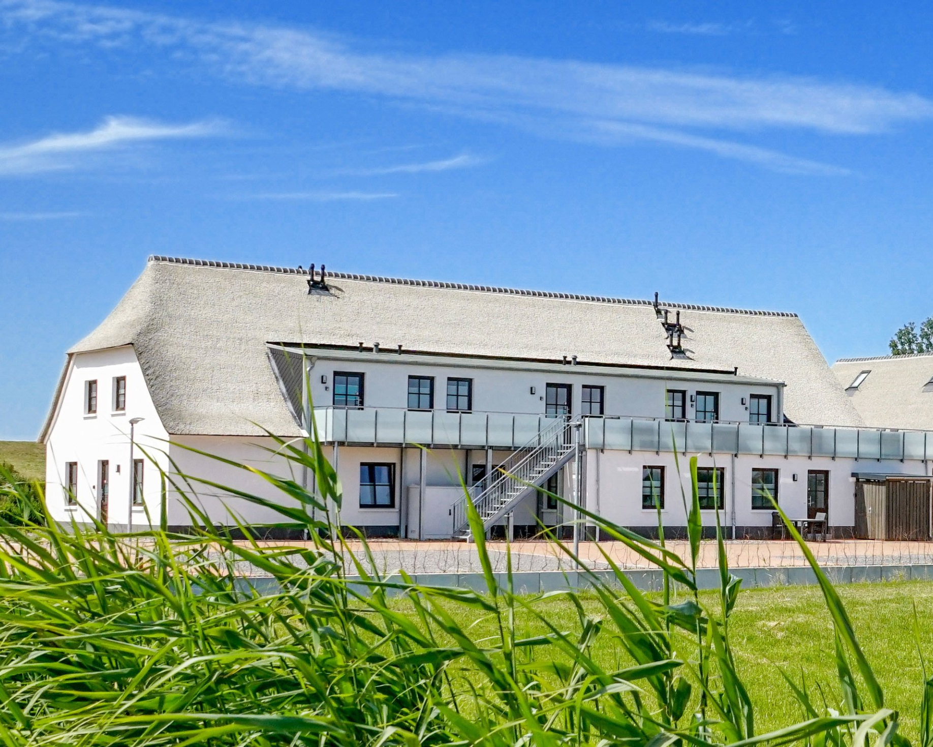 Weißes Gebäude mit Reetdach, blauer Himmel im Hintergrund. Grünes Gras im Vordergrund.