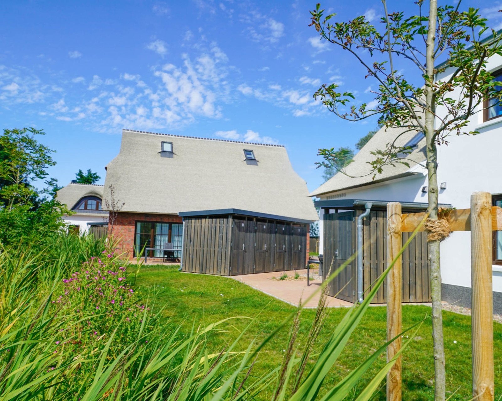 Reetgedeckte Häuser mit hölzernen Lagereinheiten und Rasenflächen unter blauem Himmel.