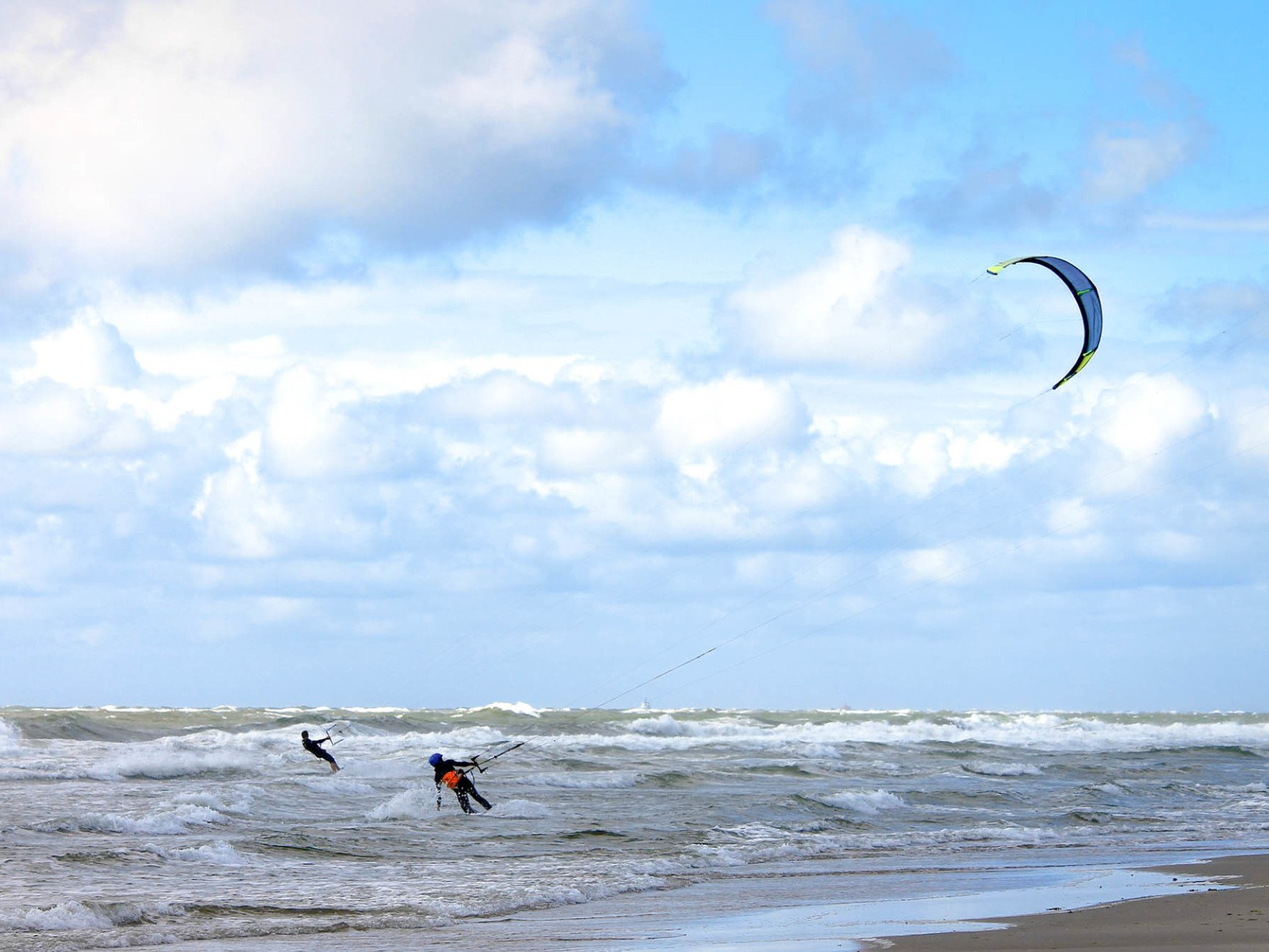 Zwei Kitesurfer auf unruhigem Wasser, ihr Kite am Himmel, an einem bewölkten Tag.