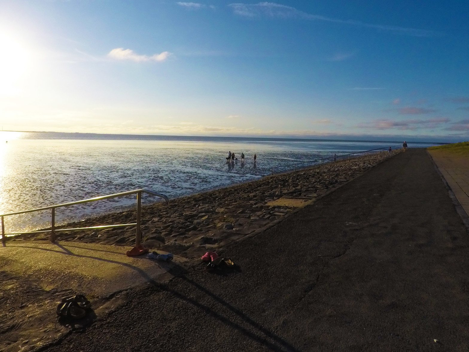 Strahlender Sonnenschein über einem Gezeitenstrand. Ein gepflasterter Gehweg säumt das Ufer.