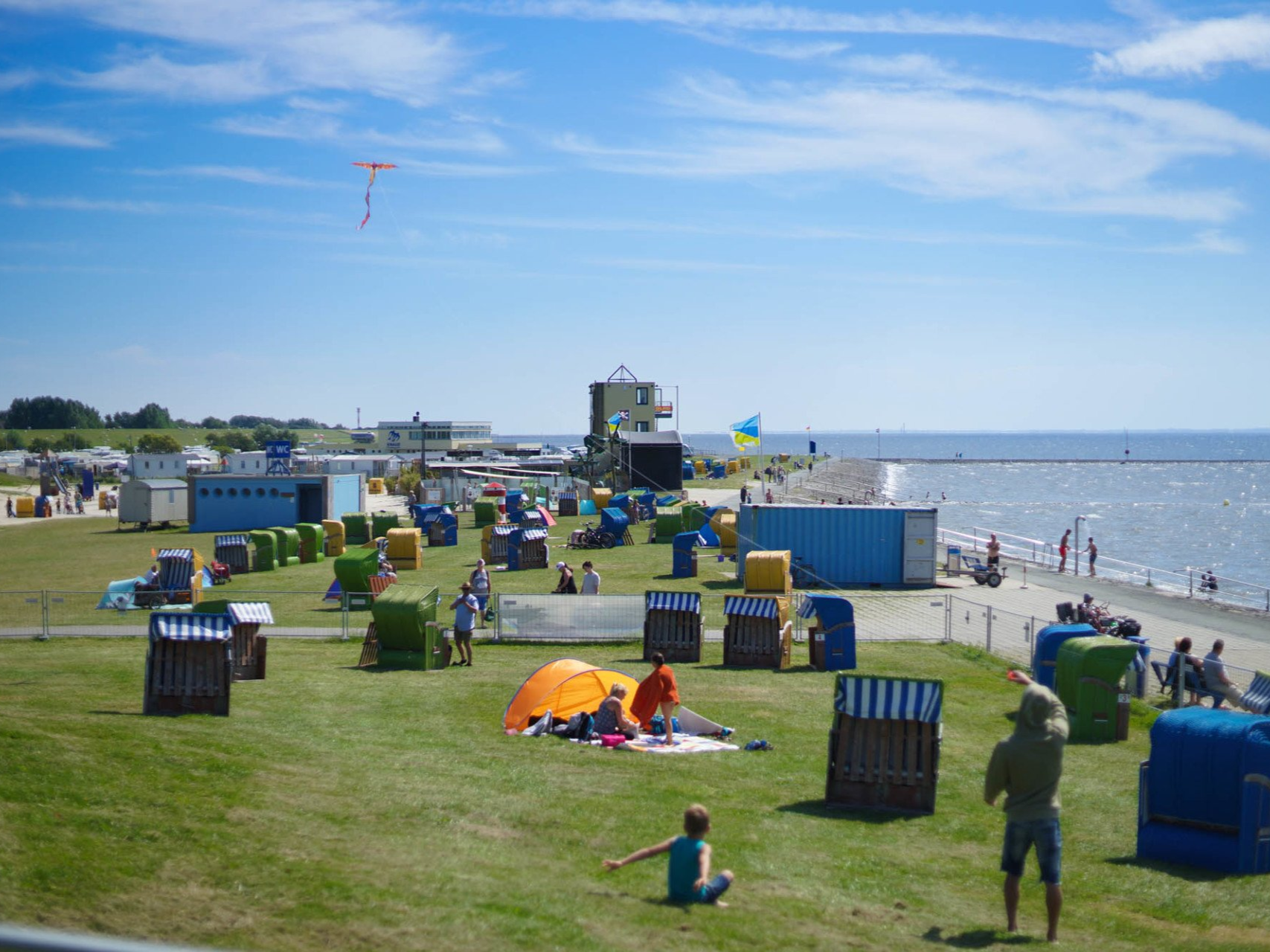 Strandszene mit Menschen, bunten Stühlen, einem fliegenden Drachen und einem blauen Meer unter sonnigem Himmel.