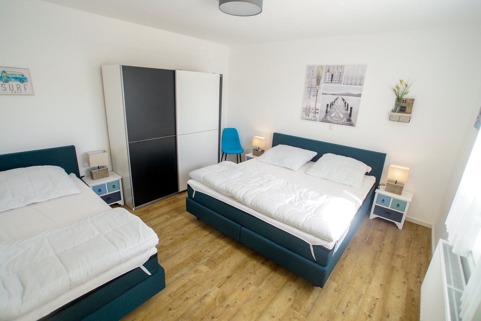 Schlafzimmer mit drei Betten, blau-weißer Farbgestaltung, Kleiderschrank und Fenster.