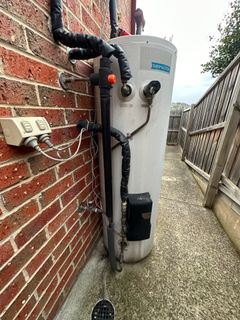 Hot water heater outside against a brick wall. Black pipes, electrical box, and a walkway.