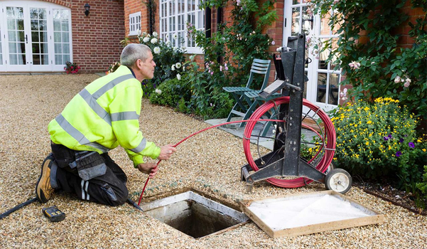 Worker in yellow vest inspecting drain with camera, kneeling by open manhole on gravel driveway.