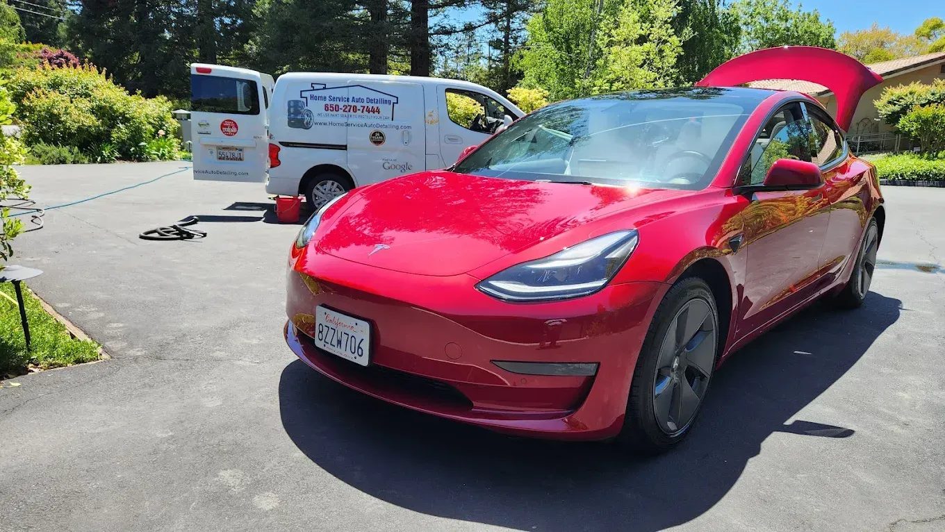 Red Tesla Model 3 car with the trunk open, parked in a driveway next to a service van.