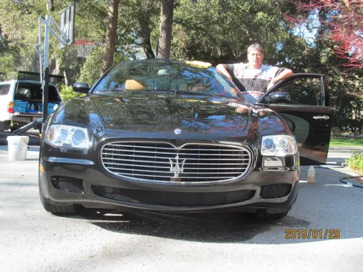 Man leaning on black Maserati in a driveway. Car is shiny, door open, trees in background.