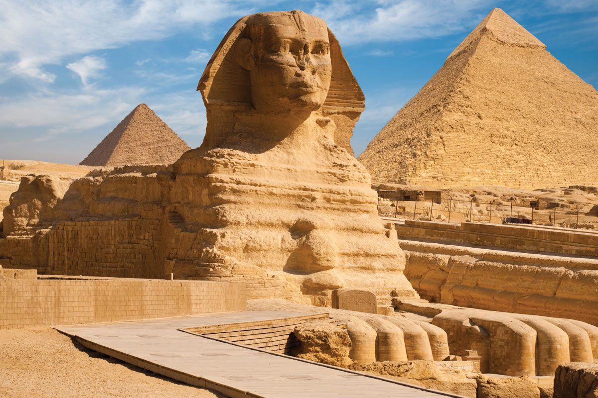 A statue of a sphinx in front of two pyramids in the desert.