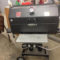 A black charcoal grill with a diamond-plate front shelf and temperature gauge sits on a wheeled stand in an indoor space.