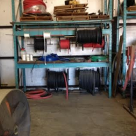 A blue metal shelving unit in a warehouse holding various spools of electrical cable, wire, and stacked wooden pallets.