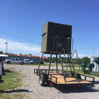 A green deer hunting blind mounted on a metal trailer with a staircase, parked on a gravel lot under a clear blue sky.