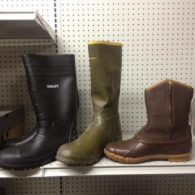 Three different styles of boots—black, olive green, and brown—sit on a white shelf against a pegboard background.