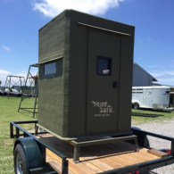 A dark green Hunt Safe hunting blind sits secured on a flatbed trailer outdoors on a sunny day.