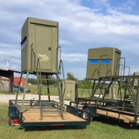 Two green, elevated hunting blinds on trailers parked in a grassy field under a cloudy sky.