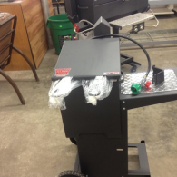 A new, black portable outdoor grill on wheels, with wrapped cooking utensils and a green-handled gas hose, in a shop.