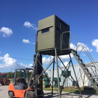 A forklift holding a green, elevated hunting blind on a metal stand outdoors under a blue sky.