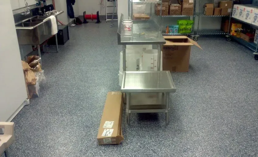 Stainless steel work tables and cardboard boxes on a speckled gray floor in a commercial kitchen.