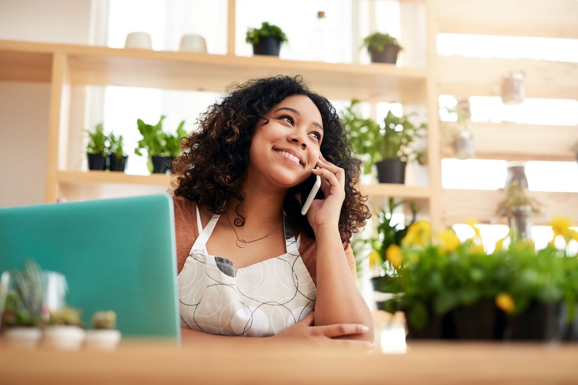 A woman is sitting at a table talking on a cell phone in front of a laptop.