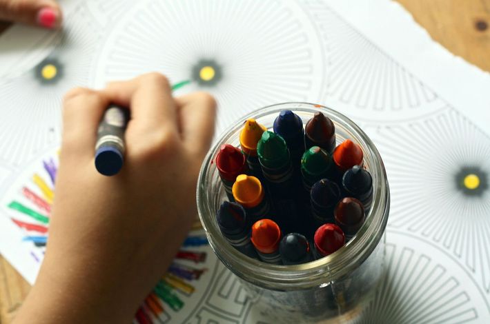 Child's hand coloring a mandala with a blue crayon, a jar of crayons nearby, on a wooden table.