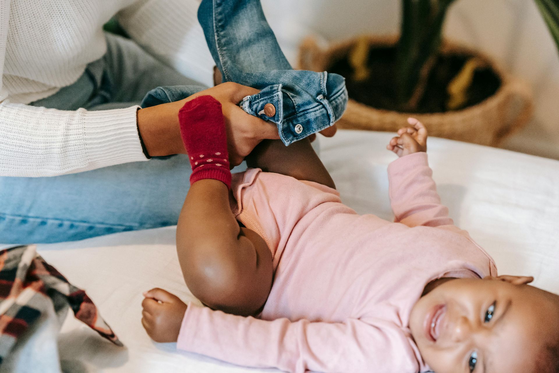 Person putting jeans on a smiling baby wearing a pink onesie, inside a home.