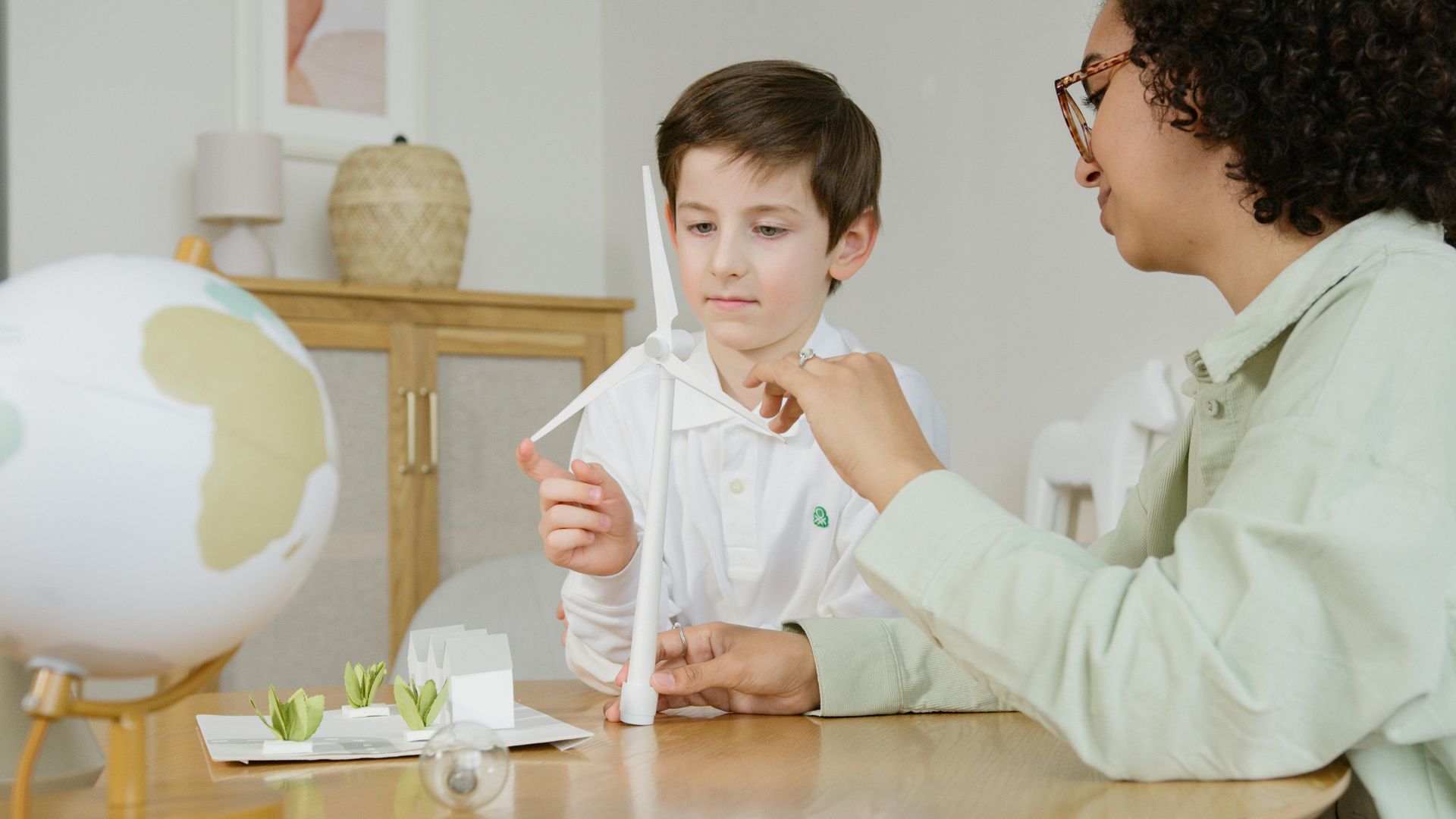 A young boy and woman examine a model wind turbine at a table; a globe and model houses are also present.
