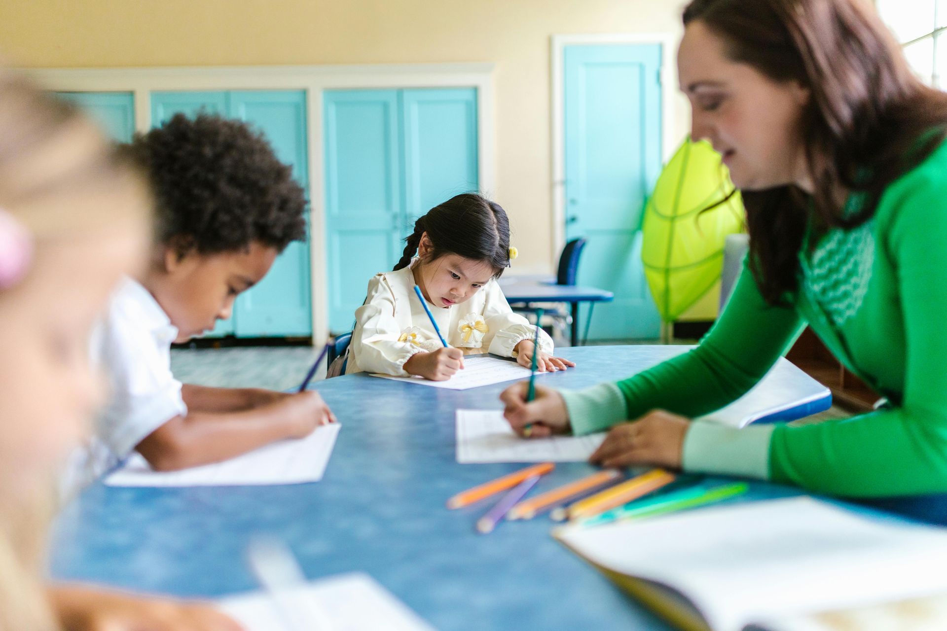 Teacher helping children write at a blue table in a classroom.