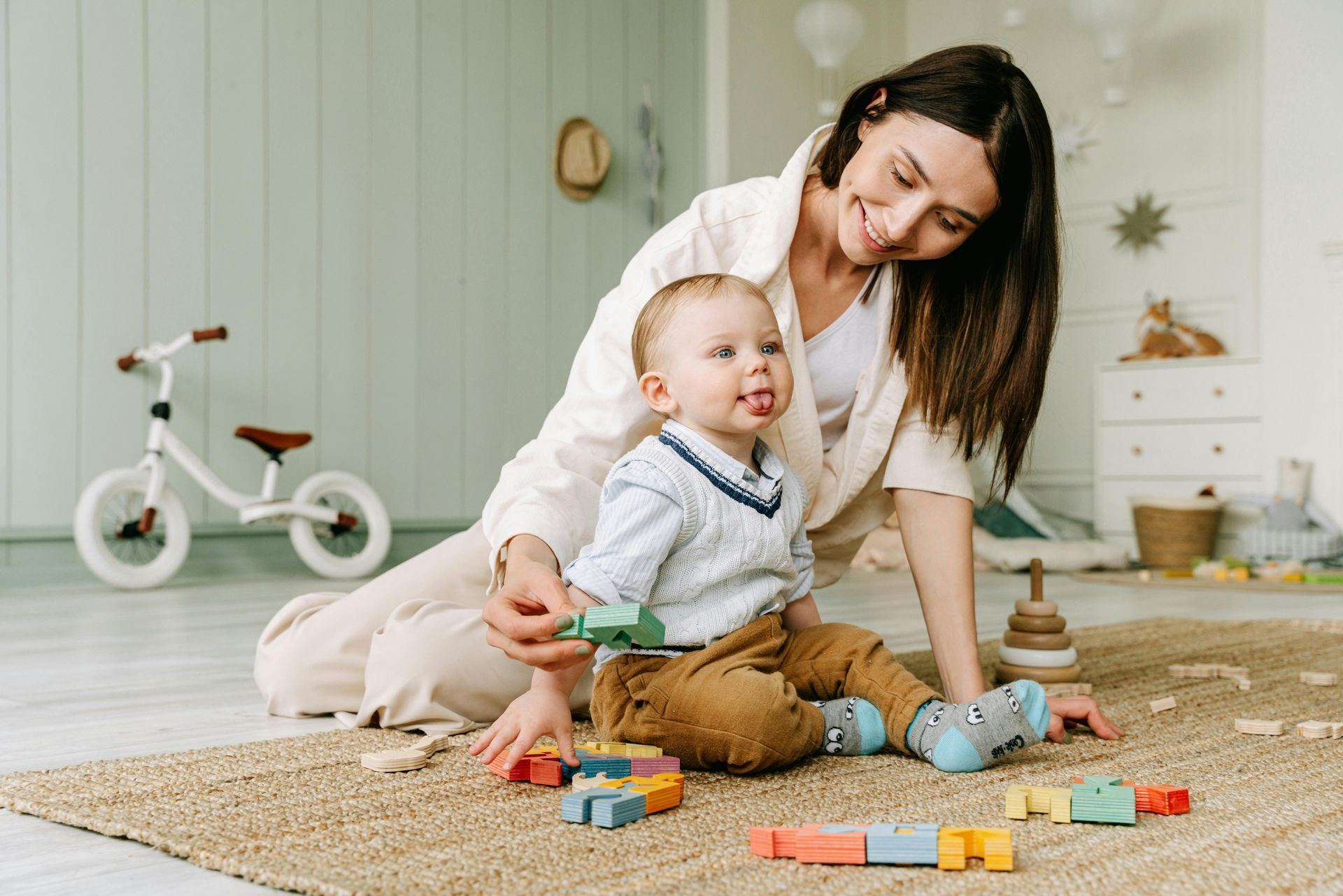 Woman smiles at toddler playing with blocks on a rug in a light-colored room.