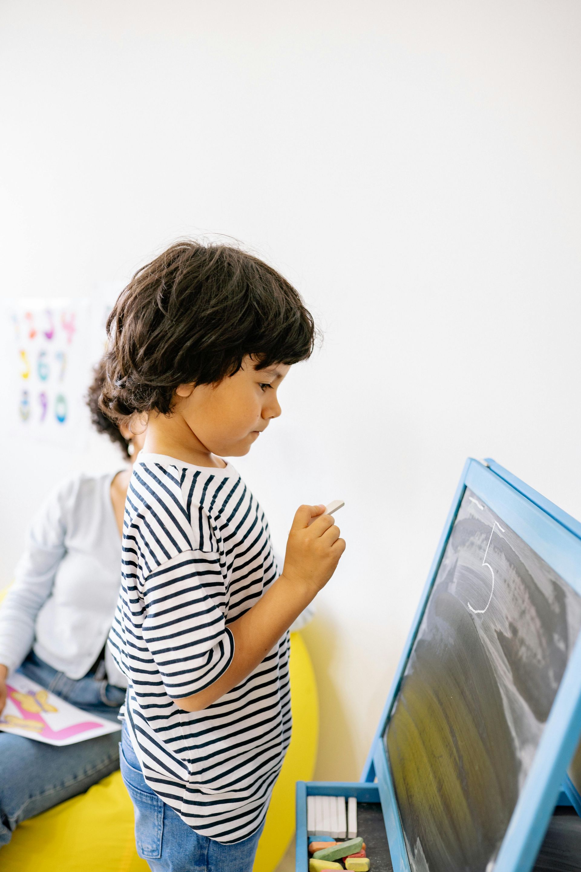 Boy drawing on a chalkboard easel, wearing striped shirt. Woman watches from behind on a yellow beanbag.