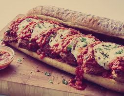 A meatball sub sandwich is sitting on a wooden cutting board next to a bowl of sauce.