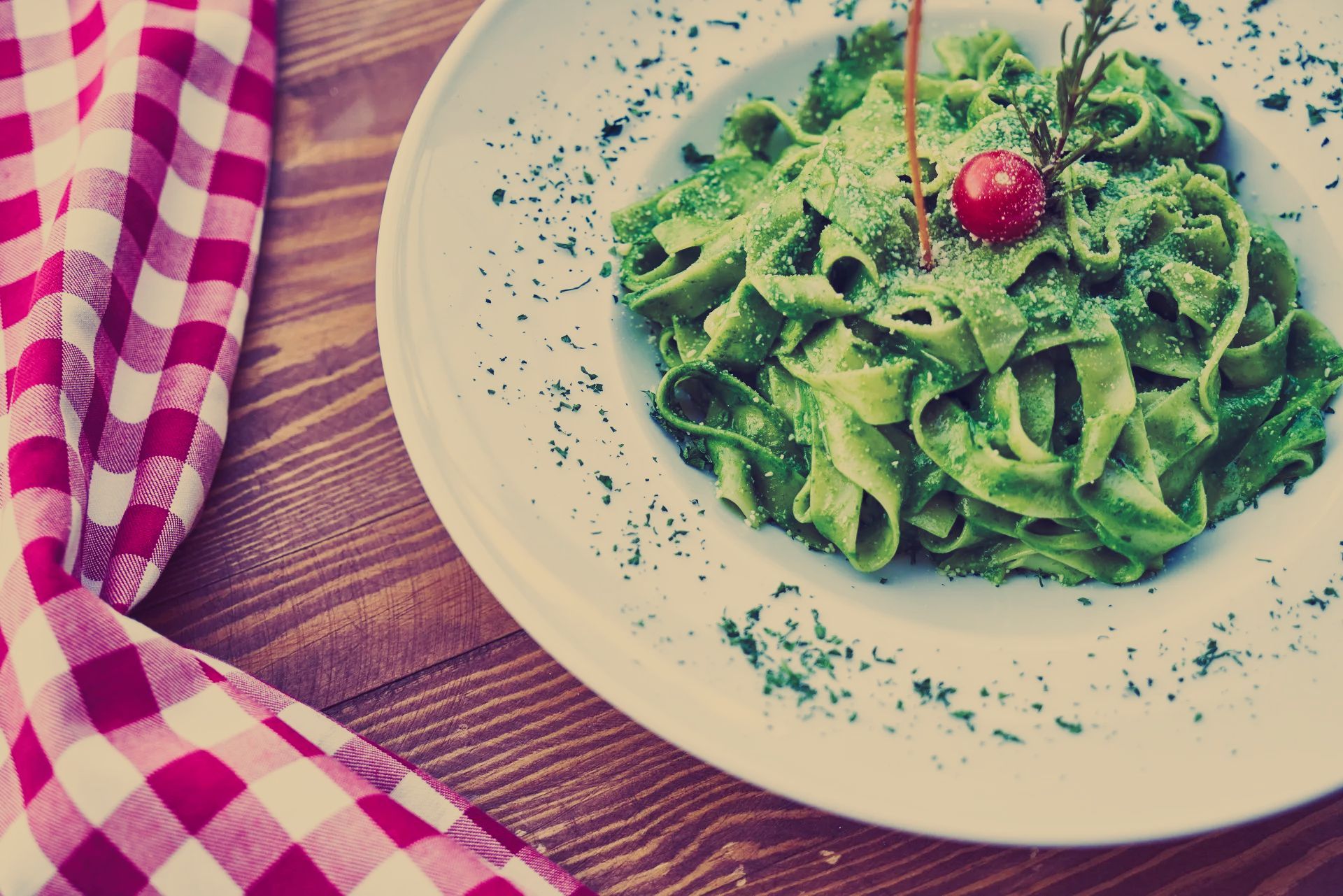 A white plate topped with green pasta and a cherry on a wooden table.