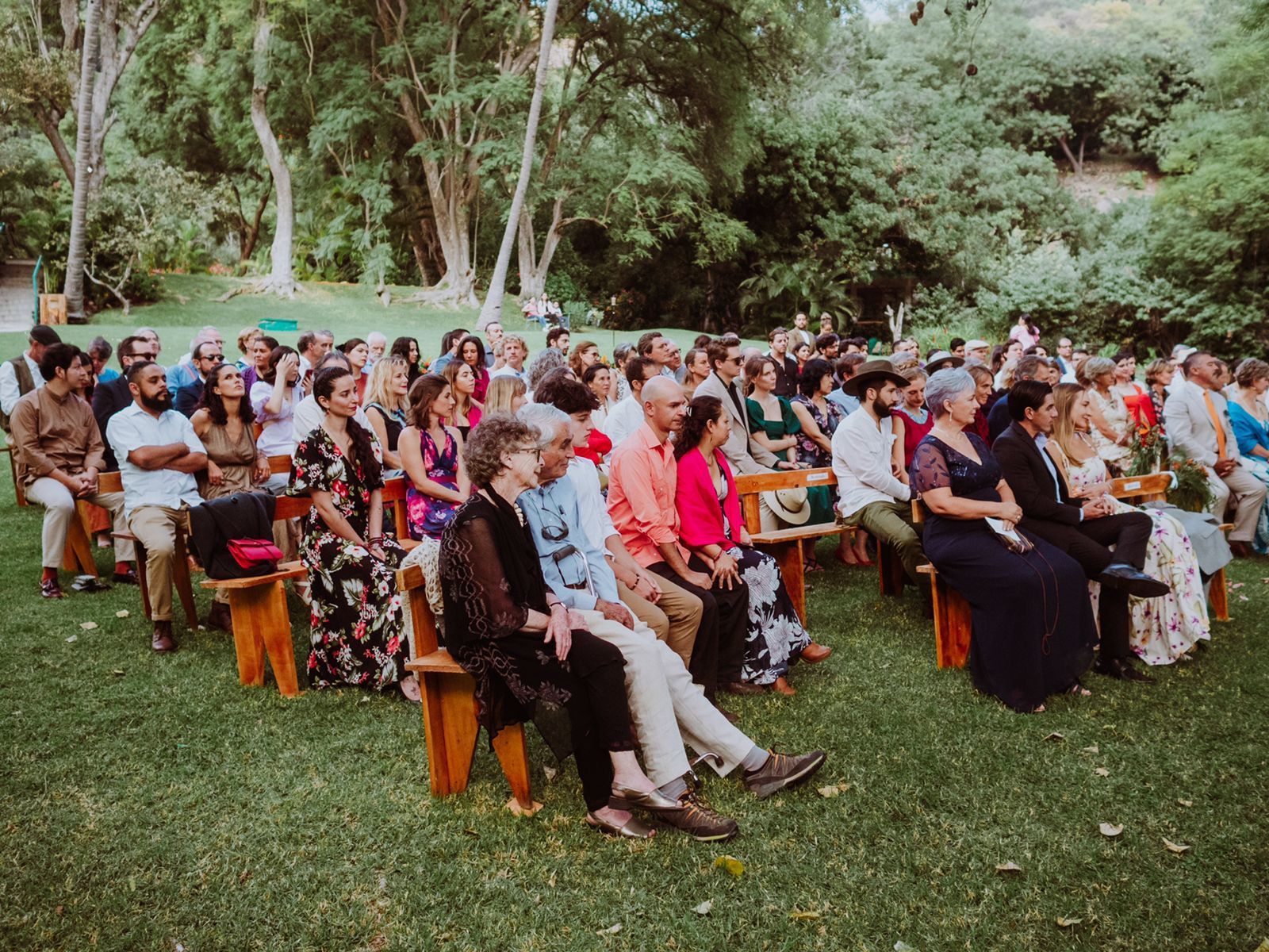 Un gran grupo de personas está sentado en bancos de madera viendo una ceremonia de boda.