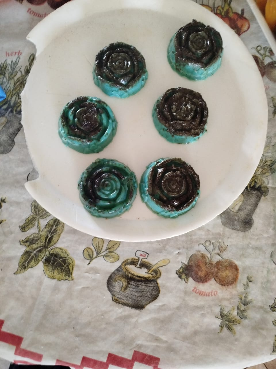 Siete caramelos en forma de rosa de color verde azulado y marrón oscuro sobre un plato blanco, colocado