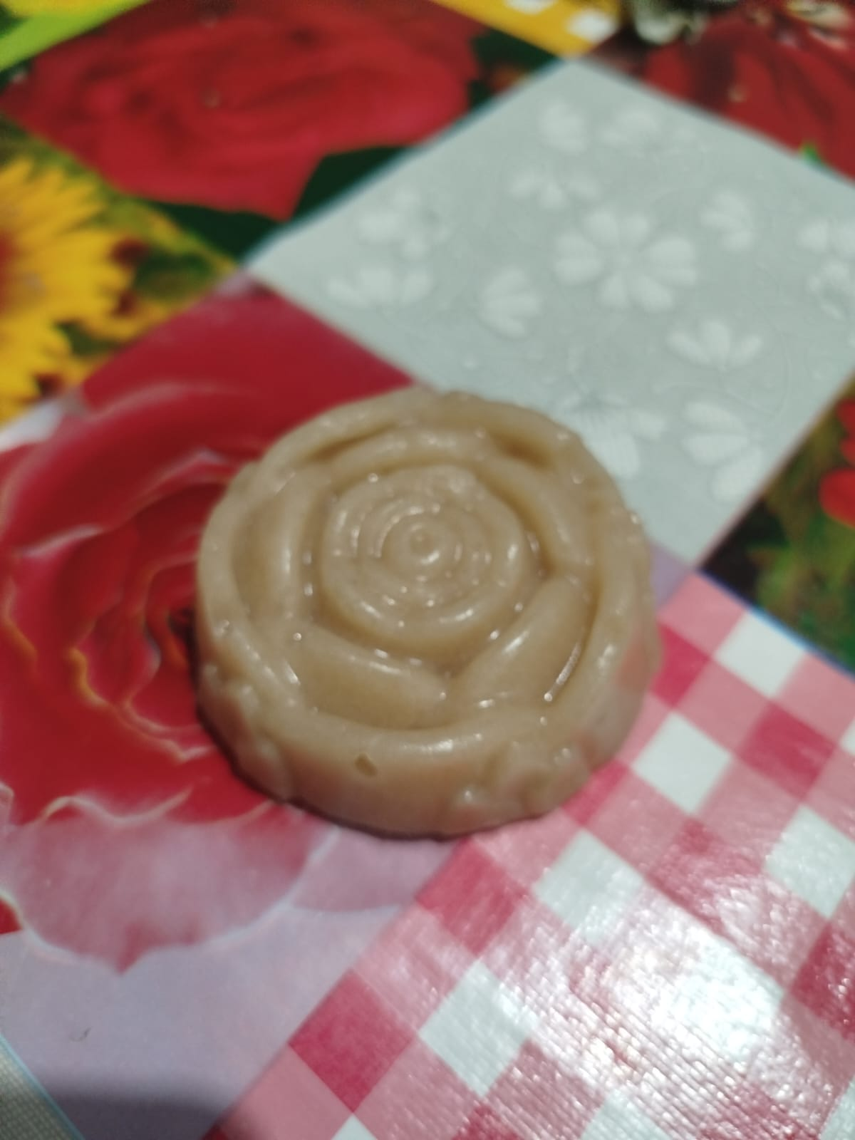 Jabón marrón claro en forma de rosa sobre una superficie estampada de rosas rojas, flores grises y cuadros vichy rosas.