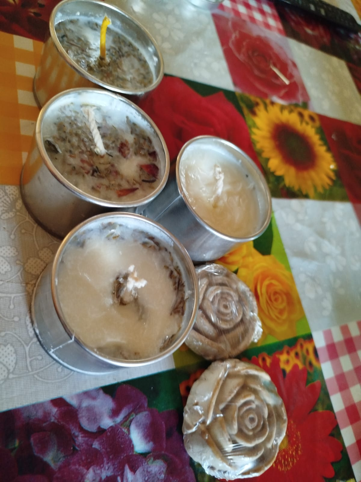 Velas en recipientes metálicos y jabones en forma de rosa sobre un mantel colorido.