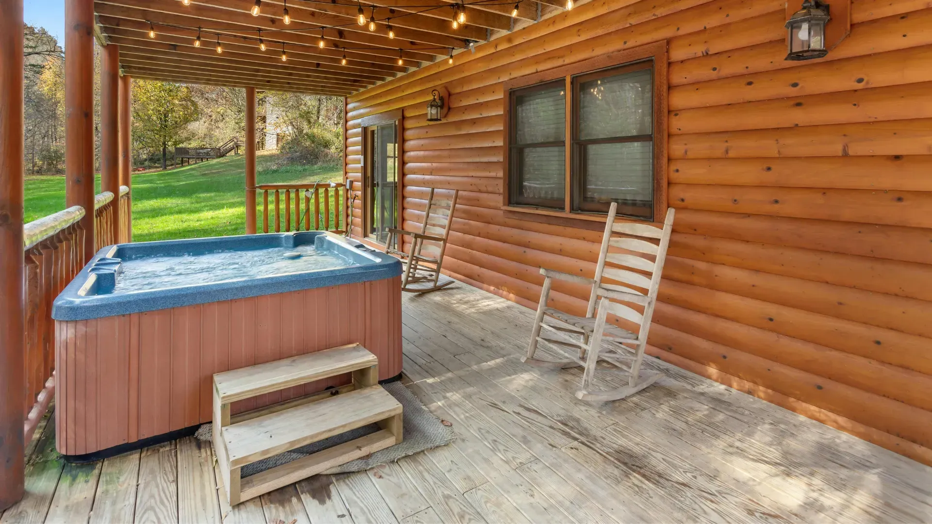 Wooden cabin porch with hot tub and rocking chairs; string lights overhead.