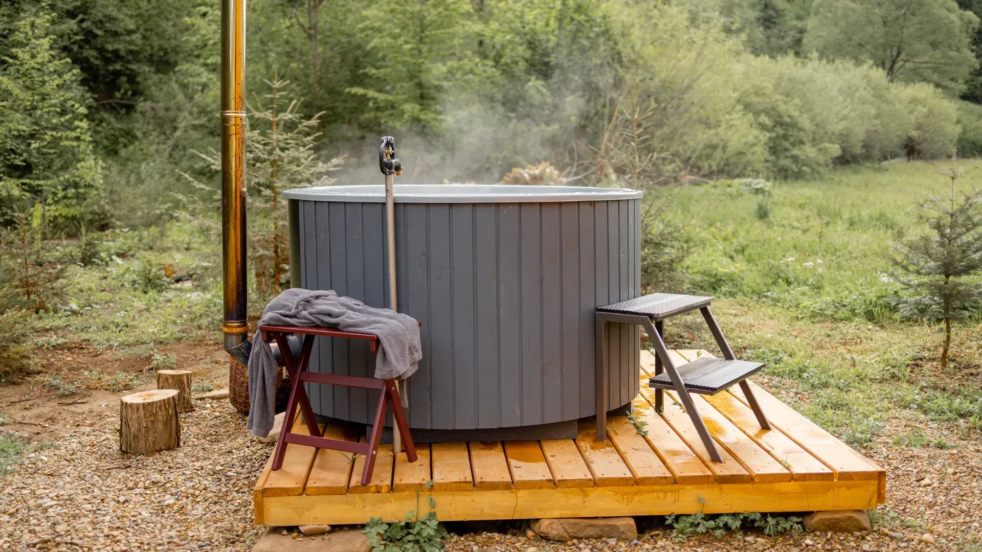 Wooden hot tub in a natural setting, with steam rising. Gray exterior, wooden steps, and towel on a stool.