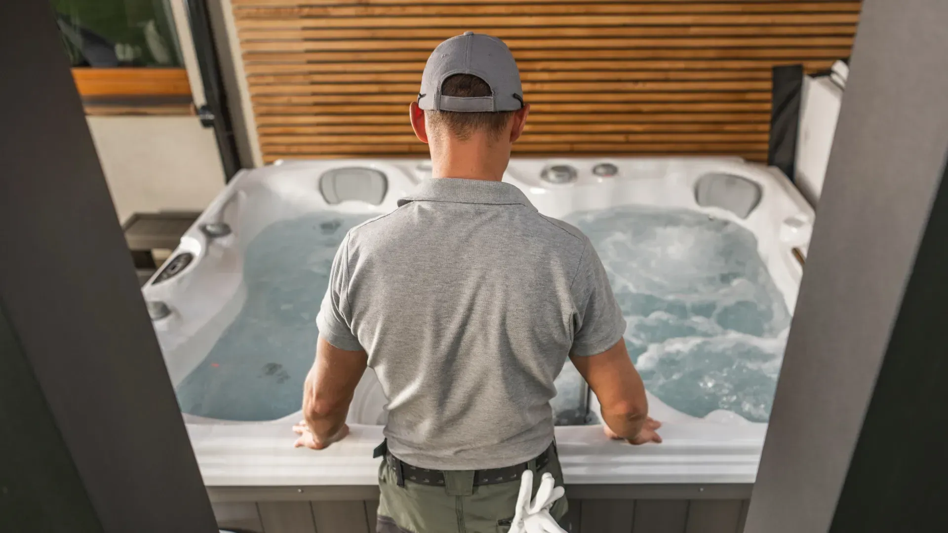 Man in gray shirt and cap stands near a hot tub outdoors, inspecting it.