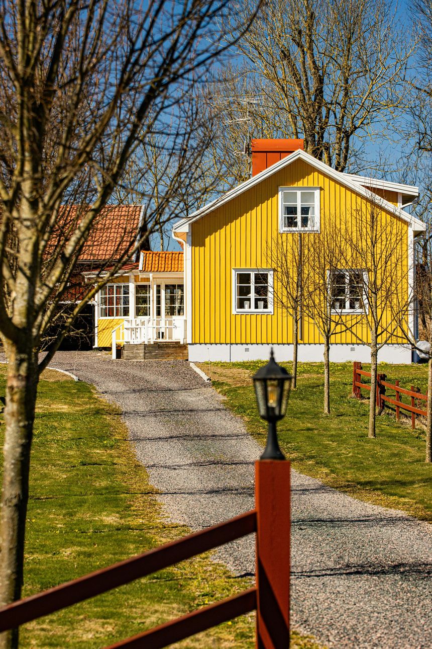 Yellow house with white trim beside a gravel path, framed by trees and a red fence in autumn.