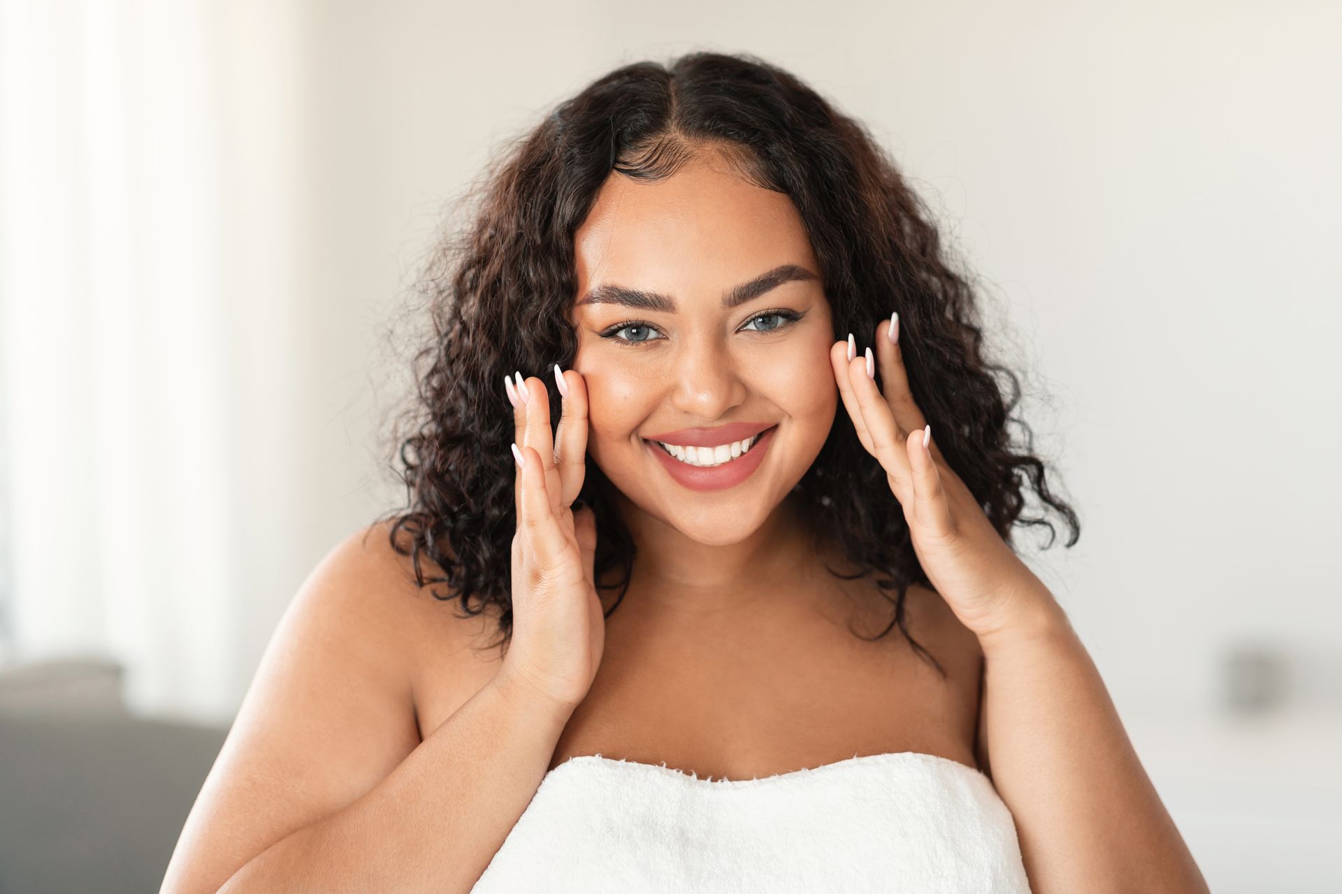 A woman wrapped in a towel is smiling and touching her face.