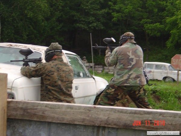 Two people in camouflage aiming paintball guns from behind a barrier beside white cars in a grassy area.