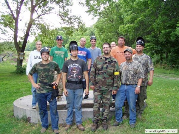 Group of people posing outdoors in camouflage and casual clothing near a concrete structure and trees