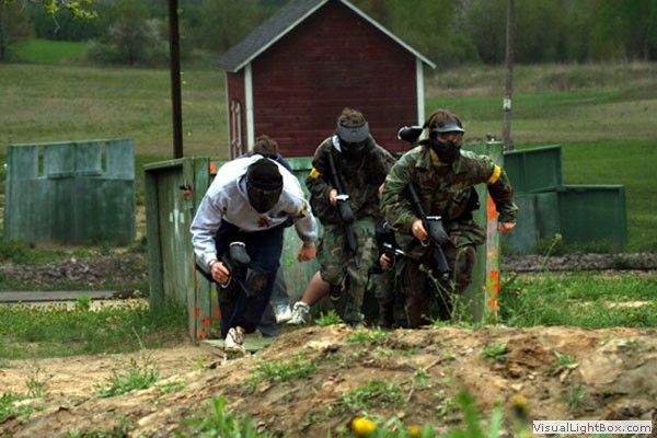 Four paintball players sprint crouched across a field toward cover near a red shed