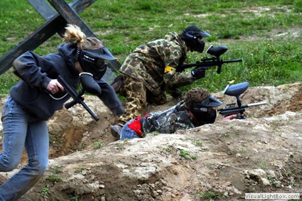 Three people in tactical gear crouch behind a dirt berm aiming paintball guns outdoors.