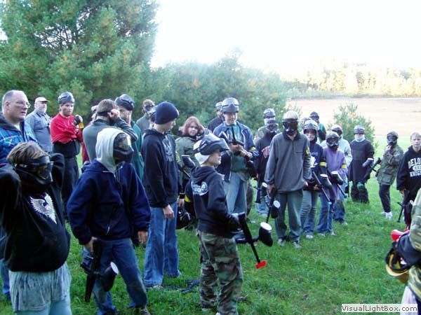 Group of people in hoodies and camouflage standing outdoors in a grassy field, gathering around a speaker.