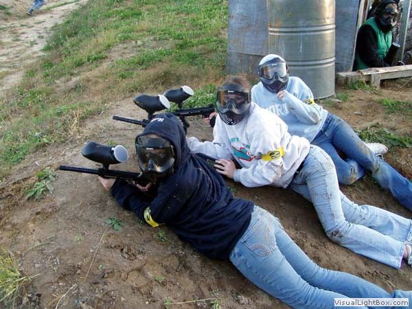 People lying prone with paintball markers behind a dirt berm, wearing masks and goggles.